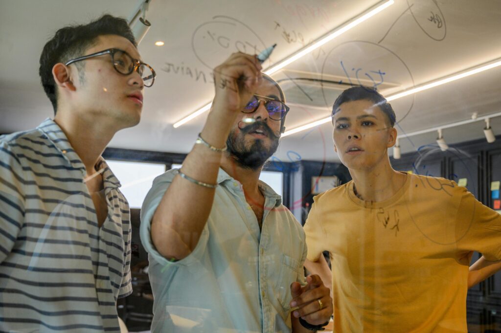 3 men in casual clothes write on a piece of glass with a whiteboard marker, focusing intensely on what they're writing
