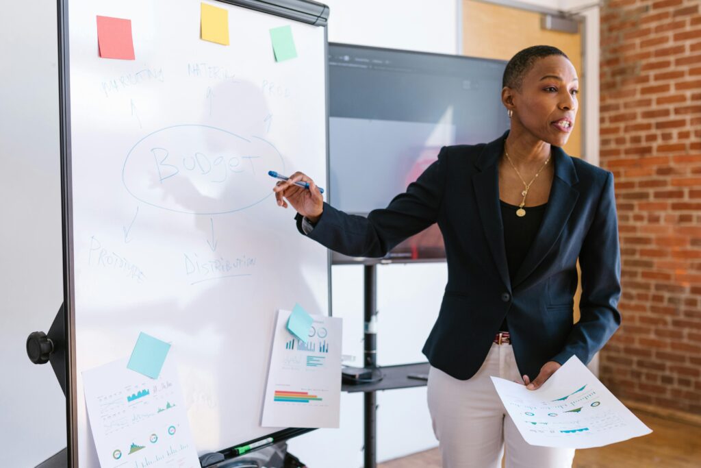 A black woman stands in front of a white board, presenting to a room